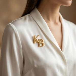 May include: A gold-colored brooch featuring the intertwined letters "KB" is pinned to a white satin garment. The brooch is in sharp focus, with the soft fabric of the garment and the background slightly blurred.