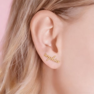 May include: Gold-colored stud earrings spelling the name "Sophia" in a cursive script. The earrings are displayed on an ear against a soft pink background, showcasing the jewelry's delicate design and personalized touch.