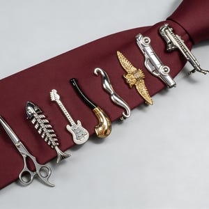 May include: An assortment of silver tie clips featuring diverse designs such as scissors, a fish skeleton, a guitar, a pipe, a wave, an eagle, a car, and a saxophone. The clips are arranged on a burgundy tie.