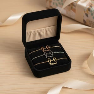 May include: Three dog-shaped bracelets in a black velvet jewelry box. Each bracelet is a different metal color: gold, rose gold, and silver. The name "Mina" is written on each bracelet. The box is open, revealing the bracelets.