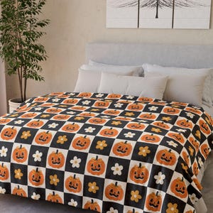 Può includere: Una coperta a tema Halloween con un motivo ripetuto di zucche arancioni, fiori bianchi e fiori gialli su uno sfondo a scacchi bianco e nero. La coperta è drappeggiata su un letto.