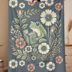 Puede incluir: Una manta azul y verde con un patrón floral y una rana leyendo un libro. La rana es verde con la barriga blanca y está sentada en un lecho de flores. Las flores son blancas, rosas y naranjas.