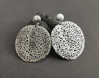 Könnte beinhalten: Ein Paar silberfarbene Ohrringe mit einem strukturierten, porösen Design. Jeder Ohrring hat einen kreisförmigen, durchbrochenen Anhänger und einen kleinen, runden Stecker. Die Ohrringe sind vor einem dunkelgrauen Hintergrund platziert.