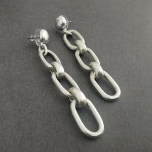 May include: A pair of silver-toned chain link earrings. Each earring features a post back and a series of oval-shaped links connected to form a dangling chain. The earrings have a matte finish and are displayed against a gray background.