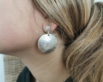 May include: Silver hammered disc earrings with a smaller silver disc on the top. The earrings are worn by a person with long blonde hair.