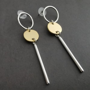 Puede incluir: Un par de pendientes colgantes plateados y dorados. Cada pendiente presenta un aro plateado, un disco dorado y una barra larga plateada. Los pendientes se muestran sobre un fondo gris oscuro.