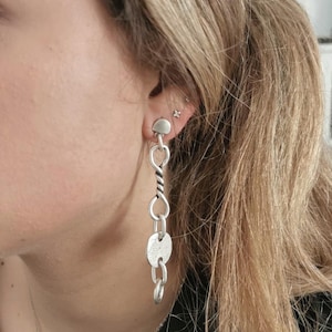 May include: Silver chain earrings with a twisted chain and a round charm. The earrings are hanging from a person's ear.