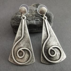 May include: A pair of silver-toned earrings with a triangular shape and a textured finish. Each earring features a curved, swirled design. The earrings are displayed on a dark gray surface.