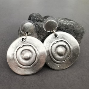 Può includere: Un paio di orecchini d'argento con un design circolare. Ogni orecchino presenta un motivo di cerchi concentrici testurizzati.