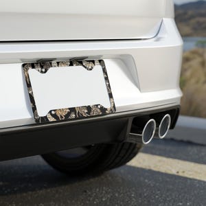 Könnte beinhalten: Ein Nummernschildrahmen mit schwarzem Hintergrund und einem sich wiederholenden Muster von Tigern. Der Rahmen ist an der Rückseite eines weißen Autos angebracht.