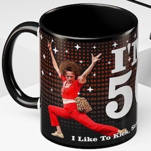 May include: Black ceramic mug featuring a woman in a red outfit with the text "I'm 50" and "I Like To Kick, Strut". The background is dark with white stars and orange dots. The mug has a black handle.