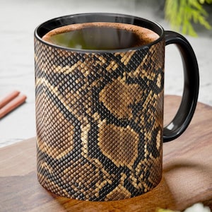 Könnte beinhalten: Schwarze Kaffeetasse mit Schlangenmuster in Braun- und Beigetönen. Die Tasse hat einen schwarzen Henkel und Rand und ist mit einer dunklen Flüssigkeit gefüllt. Sie steht auf einer Holzoberfläche.
