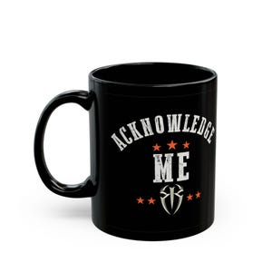 May include: Black ceramic mug with the words "ACKNOWLEDGE ME" in white, along with orange stars and a silver emblem. The mug has a curved handle and a glossy finish.