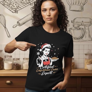 May include: Black t-shirt featuring a retro illustration of a woman in a Santa hat and apron holding cookies. The text reads "Certified Gingerbread Cookie Expert". The background includes baking tools.