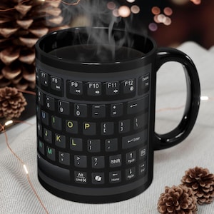 Peut inclure: Mug en céramique noire avec un motif de clavier, avec des touches comme F7, F8 et Shift. La tasse a une anse noire et contient une boisson fumante. La tasse est entourée de pommes de pin et de guirlandes lumineuses.