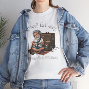 May include: White t-shirt featuring a cartoon illustration of a man playing a guitar, with the text "What A Long, Strange Trip It's Been." The illustration includes an amplifier and a guitar case. The shirt is worn by a person wearing a denim jacket.
