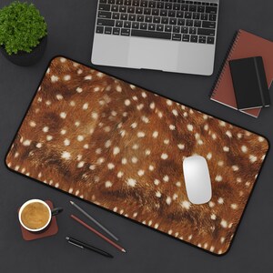 May include: A rectangular desk mat featuring a brown and white spotted pattern, resembling deer fur. A white computer mouse sits on the mat, with a laptop, notebooks, and a cup of coffee visible in the background.