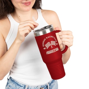 May include: A red travel mug with a silver lid and a black handle. The mug features the text "SUNDAY RACING CLUB" and "FORMULA ONE" with checkered flags. The mug is held by a person wearing a white tank top and blue jeans.
