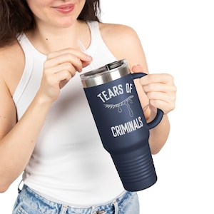 May include: A navy blue travel mug with a silver lid and handle. The mug features the text "TEARS OF CRIMINALS" in white, along with a graphic of an eye with teardrops. The mug has a clear straw.