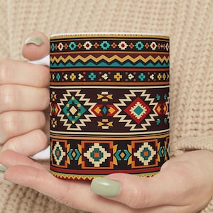 May include: A white ceramic mug with a Southwestern-inspired geometric pattern in shades of brown, teal, orange, and yellow. The design features repeating diamond and zigzag motifs, creating a vibrant and eye-catching aesthetic. The mug is held in a person's hands.