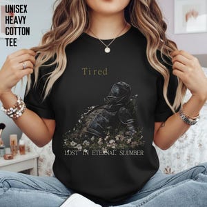 Op de afbeelding: Zwart unisex zwaar katoenen T-shirt met de woorden "Tired" en "Lost in Eternal Slumber". Het shirt heeft een afbeelding van een ridderharnas bedekt met bloemen.