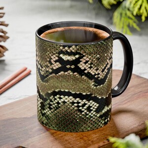 Op de afbeelding: Een zwarte koffiemok met een slangenhuiddesign in groen, bruin en zwart. De mok heeft een zwart handvat en rand en is gevuld met een donkere vloeistof. Stoom stijgt op van de bovenkant van de mok.
