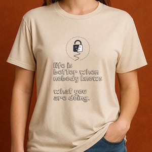 May include: Beige t-shirt with the text "life is better when nobody knows what you are doing." The shirt features a graphic of a smiling lock inside a dotted circle.