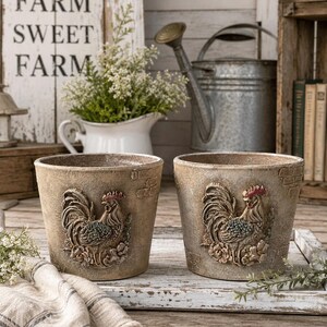May include: Two round, beige-toned planters featuring a raised, three-dimensional rooster relief on the front. The pots have a weathered, textured finish with subtle brick-like patterns embossed on the sides and a wide, open top rim.