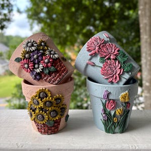 May include: Four flower pots with textured surfaces. Two pots are light pink with relief designs of sunflowers and mixed wildflowers in woven baskets. Two pots are light blue with relief designs of pink chrysanthemums and colorful tulips.