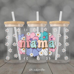 May include: Three clear glass tumblers with wooden lids and white straws. Each tumbler has a floral design with the word "mama" in the center. White daisy flowers are scattered across the glass.