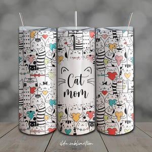 May include: A white tumbler with a lid and straw, featuring a repeating pattern of cartoon cats, hearts, and bows in various colors. The center of the tumbler has the words "Cat Mom" with cat whiskers and ears.