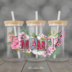 May include: Three clear glass tumblers with wooden lids and white straws. Each tumbler features the word "MAMA" in colorful block letters, surrounded by floral designs in pink, green, and white. The tumblers are on a wooden surface.