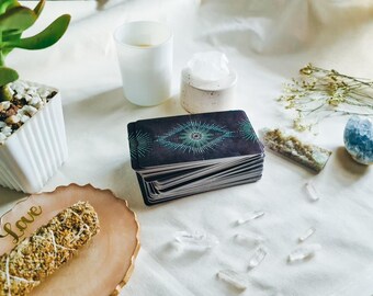 May include: A stack of tarot cards with a teal and black design, alongside crystals, a candle, and a sage bundle on a white surface. The sage bundle rests on a pink plate with the word "Love" in gold lettering.