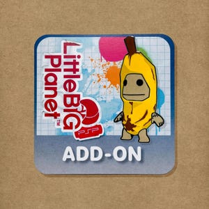 Puede incluir: Una pegatina cuadrada con el logotipo de LittleBigPlanet PSP en texto rojo, un personaje amarillo con disfraz de plátano y un fondo de cuadrícula azul con salpicaduras de pintura. La palabra ADD-ON está impresa en letras blancas en la base.