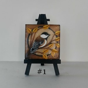 May include: A small, square painting of a bird perched on a branch with yellow leaves, set on a miniature black easel. The bird has a black and white head and a brown body. The painting is labeled with the number "#1".