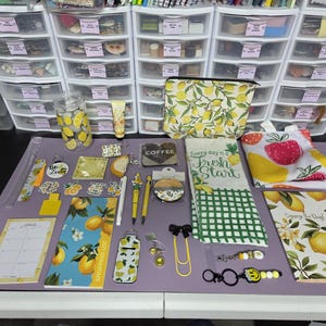 May include: A collection of stationery and accessories with a lemon theme. Items include a cosmetic bag, pens, a notepad, a tumbler, and keychains. The color palette is yellow, white, and green, with a purple mat in the background.