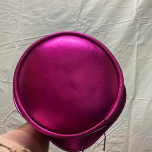 Pode incluir: Um chap&eacute;u f&uacute;csia met&aacute;lico com uma copa arredondada e uma aba. O chap&eacute;u tem uma textura lisa e brilhante e parece ser feito de um material sint&eacute;tico. Duas cordas finas e combinando pendem da lateral do chap&eacute;u.