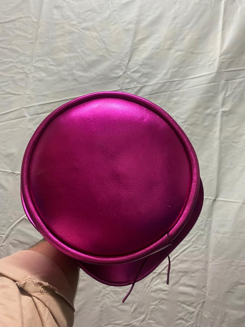 Pode incluir: Um chap&eacute;u f&uacute;csia met&aacute;lico com uma copa arredondada e uma aba. O chap&eacute;u tem uma textura lisa e brilhante e parece ser feito de um material sint&eacute;tico. Duas cordas finas e combinando pendem da lateral do chap&eacute;u.