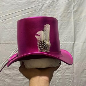 Pode incluir: Um chap&eacute;u de copa magenta com aba larga e uma pena decorativa. O chap&eacute;u tem um acabamento brilhante e uma forma cl&aacute;ssica. A pena &eacute; branca com manchas pretas.