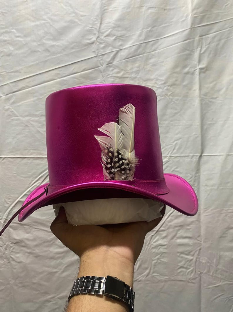 Pode incluir: Um chap&eacute;u de copa magenta com aba larga e uma pena decorativa. O chap&eacute;u tem um acabamento brilhante e uma forma cl&aacute;ssica. A pena &eacute; branca com manchas pretas.
