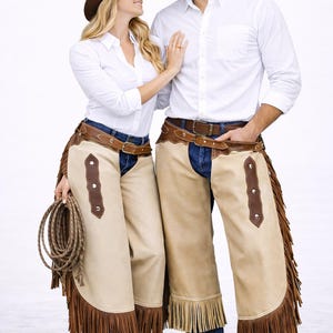 May include: A couple in Western attire, wearing brown cowboy hats, white button-down shirts, blue jeans, and tan chaps with brown fringe. One holds a rope. The chaps have brown leather accents and are secured with brown belts.