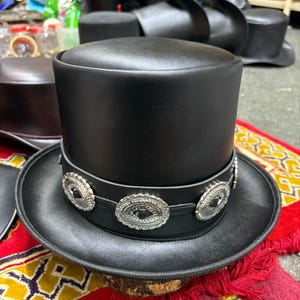 May include: A black leather top hat with a wide brim. The hat has a decorative band with silver-coloured circular embellishments. The hat is displayed on a colourful rug with red and yellow patterns, with other hats in the background.