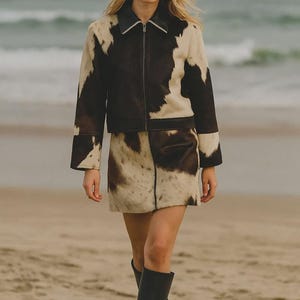 Op de afbeelding: Een tweedelige koeienhuid outfit, bestaande uit een jasje en een minirok. Het jasje heeft een zwarte kraag en ritssluiting, met een zwart-wit koeienhuid patroon. De rok heeft een bijpassend patroon. De outfit wordt gecombineerd met zwarte kniehoge laarzen.