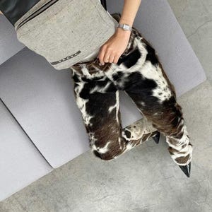 Peut inclure: Pantalon &agrave; motif de peau de vache avec des talons noirs &agrave; bout pointu. Un sac banane Prada gris est port&eacute; en travers de la poitrine. Une montre argent&eacute;e est au poignet. La personne est assise sur un canap&eacute; gris.