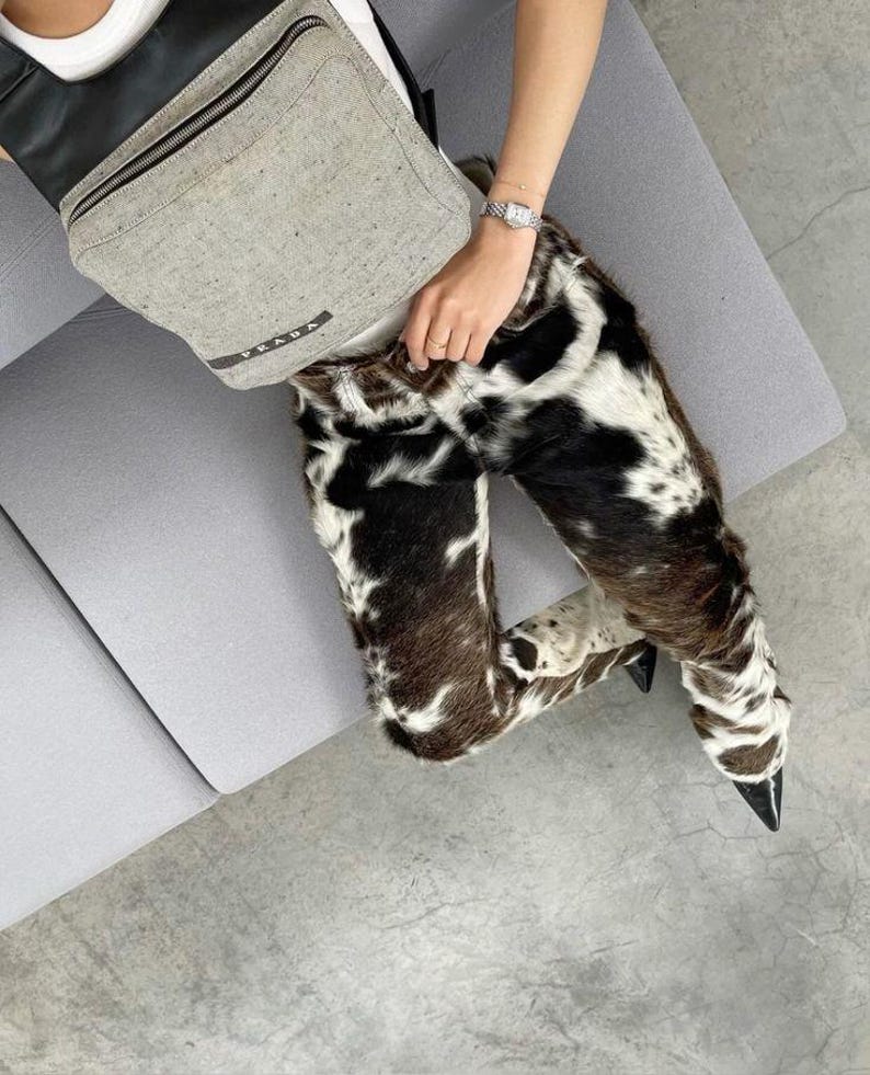 Peut inclure: Pantalon &agrave; motif de peau de vache avec des talons noirs &agrave; bout pointu. Un sac banane Prada gris est port&eacute; en travers de la poitrine. Une montre argent&eacute;e est au poignet. La personne est assise sur un canap&eacute; gris.