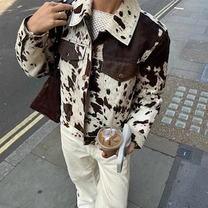 May include: A cowhide print jacket with brown and white spots. The jacket has two front pockets and a collar. The person is wearing a white sweater and cream-colored pants. A brown purse is over the shoulder.
