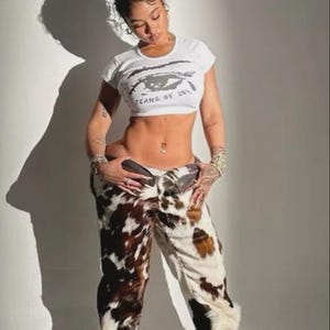 Peut inclure: Un crop top blanc avec le texte "TEARS OF JOY" et un motif d'&oelig;il. Le mod&egrave;le porte un pantalon &agrave; imprim&eacute; vache avec des taches marron et blanches. Le pantalon a une coupe &eacute;vas&eacute;e.