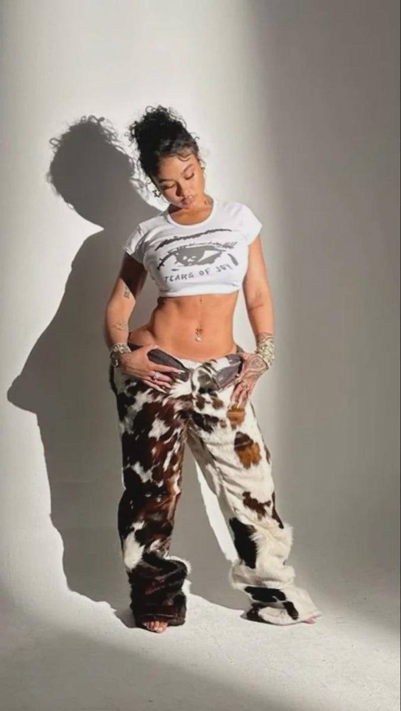 Peut inclure: Un crop top blanc avec le texte "TEARS OF JOY" et un motif d'&oelig;il. Le mod&egrave;le porte un pantalon &agrave; imprim&eacute; vache avec des taches marron et blanches. Le pantalon a une coupe &eacute;vas&eacute;e.
