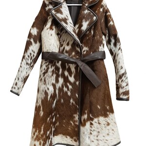May include: A long coat with a brown and white spotted pattern, hanging on a white hanger. The coat has a dark brown leather belt and trim. The coat is made of a cowhide material.