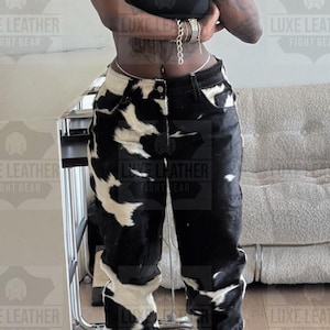 May include: Black and white cowhide-patterned jeans. The jeans have a high waist and a straight leg cut. The model is wearing a black crop top and silver jewelry. The jeans are the focal point of the image.
